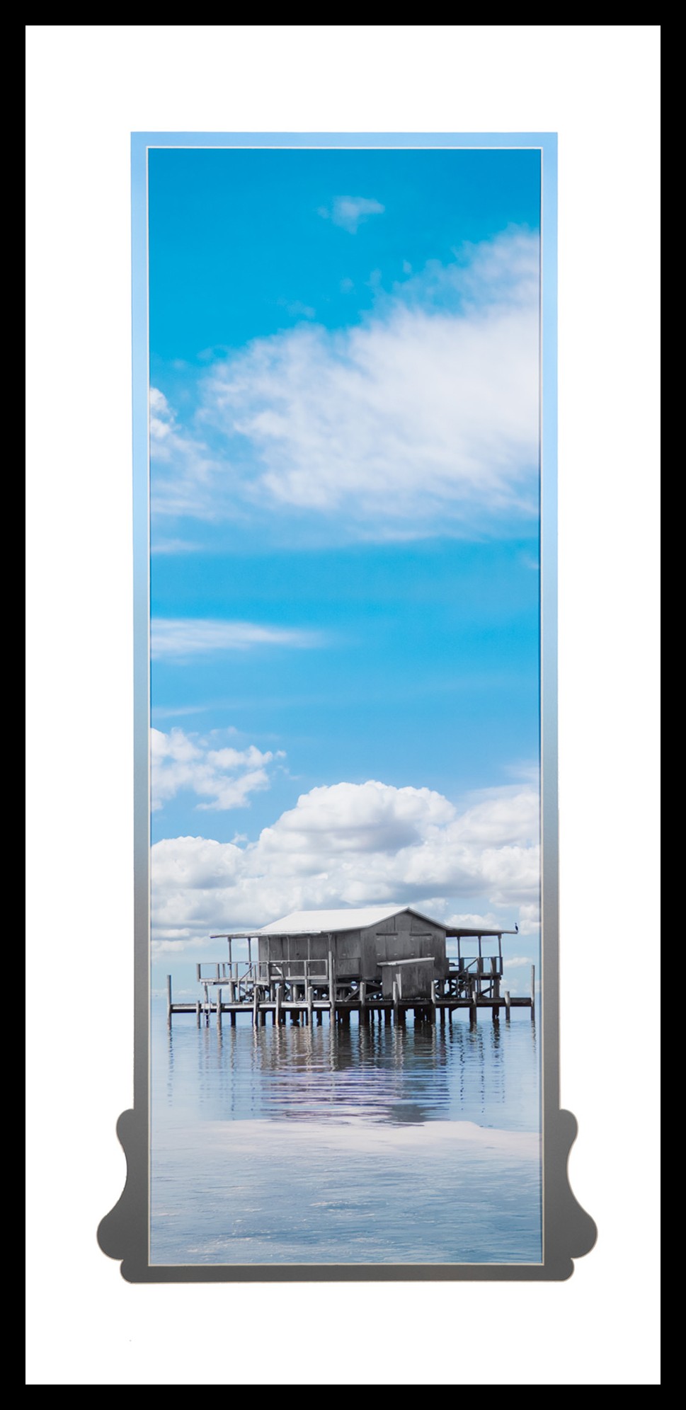 Framed photograph of a stilt house over calm water beneath a blue sky